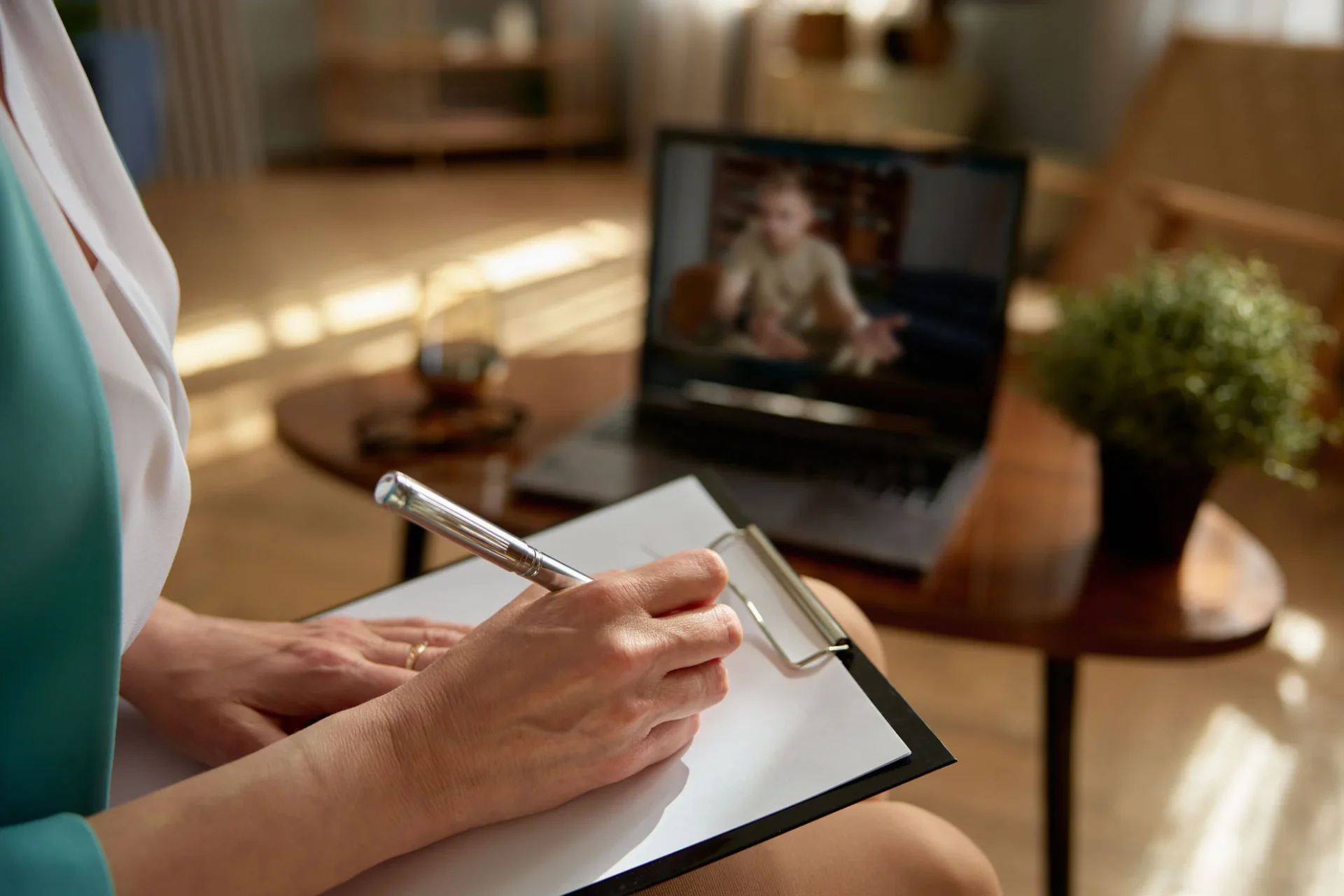 Therapist taking handwritten notes during an online couples session shown on a laptop screen