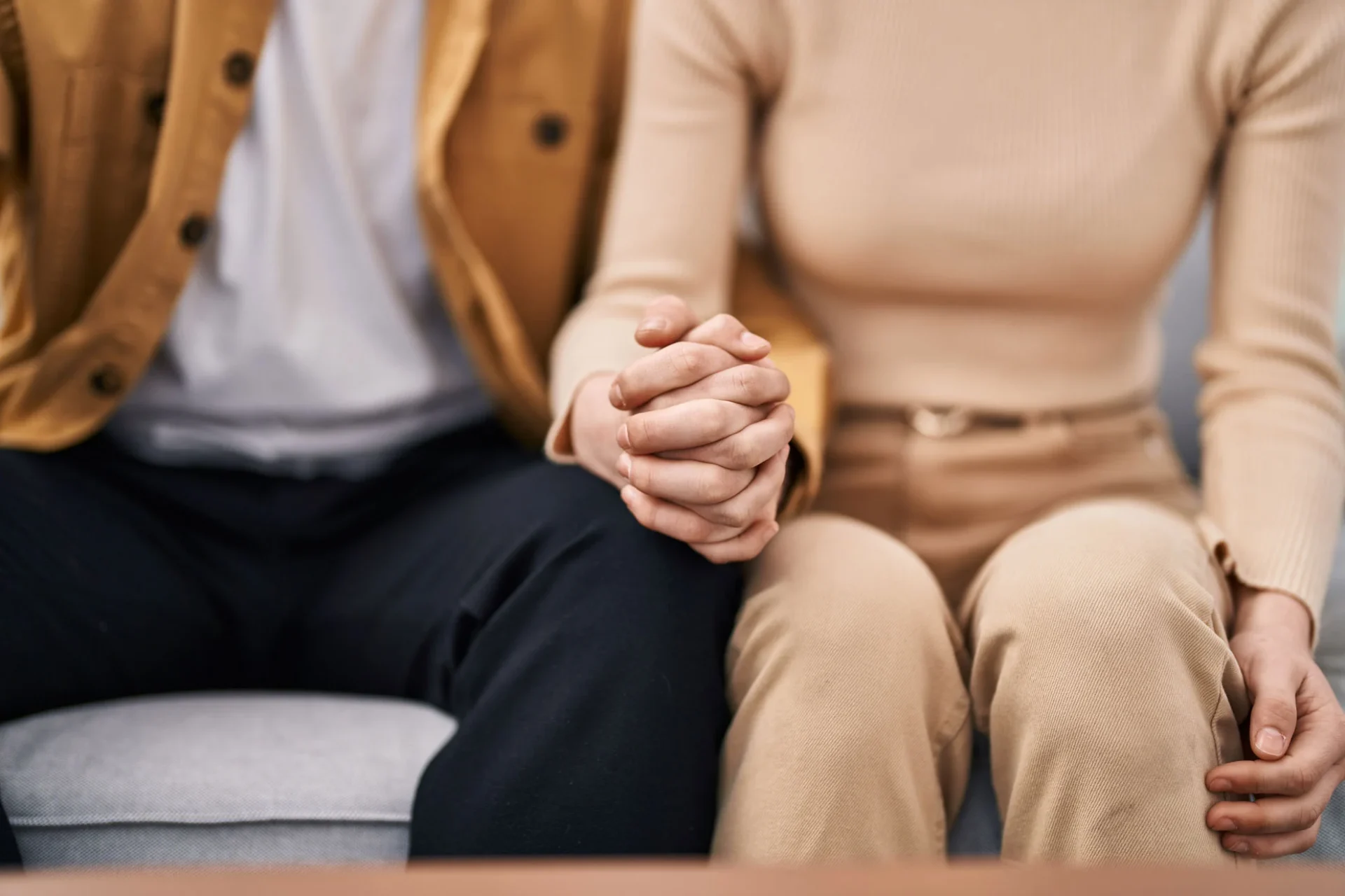 Close-up of a couple sitting side by side on a sofa, one partner's hands clasped in their lap