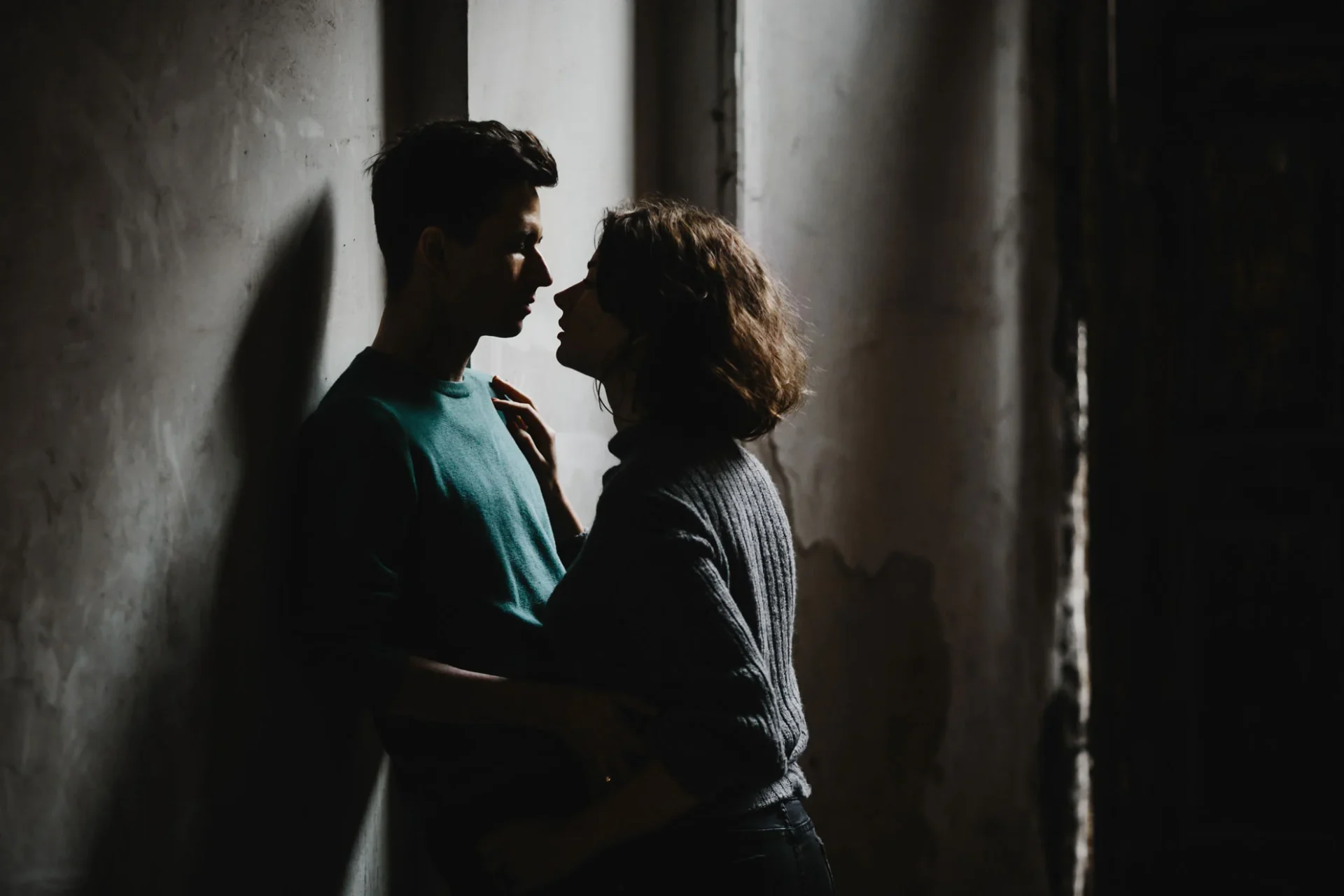 Couple standing close against a dim wall, faces lit by a narrow window