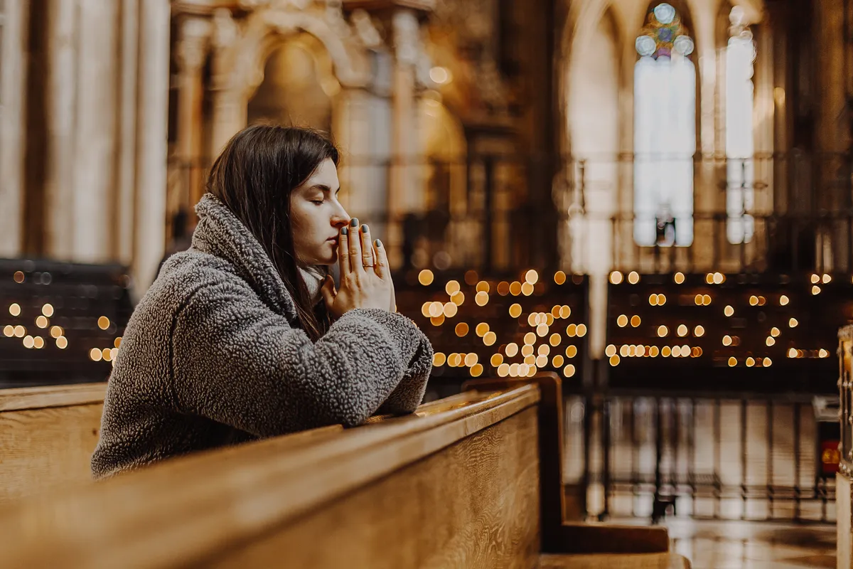 Young woman praying in a church pew with candles glowing behind her