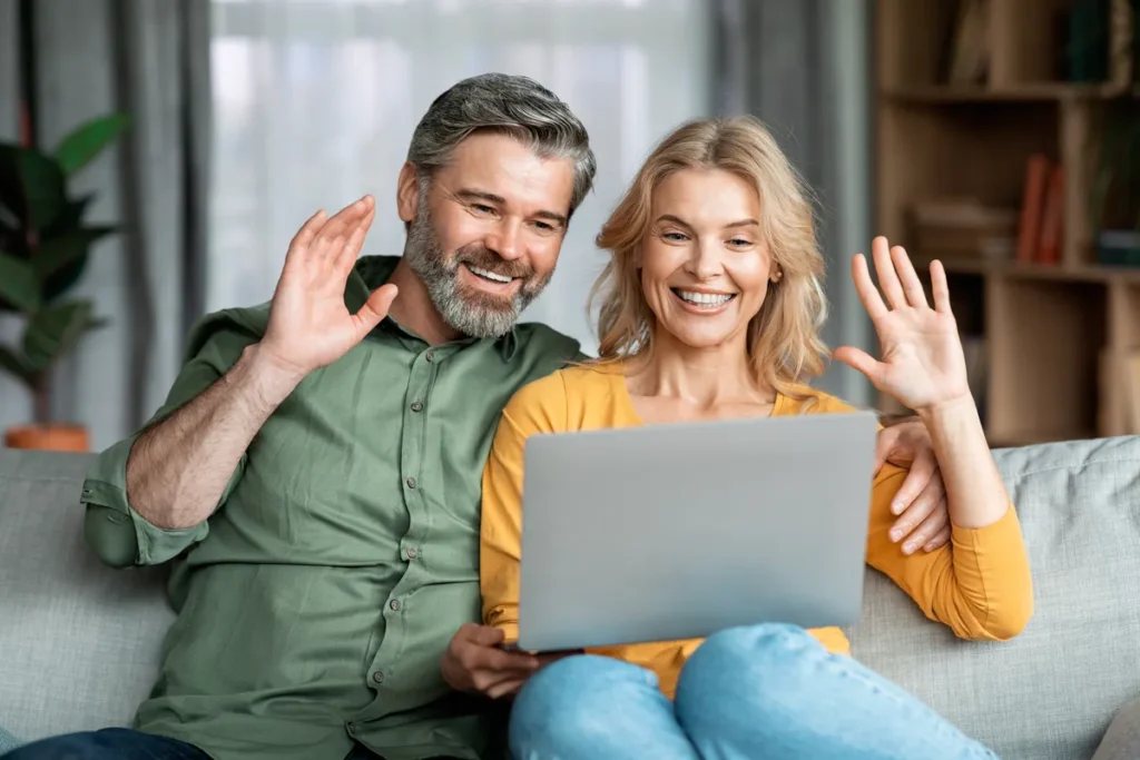 Couple waving at a video call on a laptop from their sofa