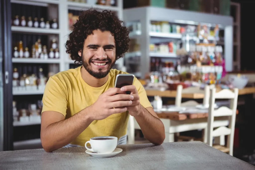 Man smiling at his phone over a coffee in a cafe while chatting online