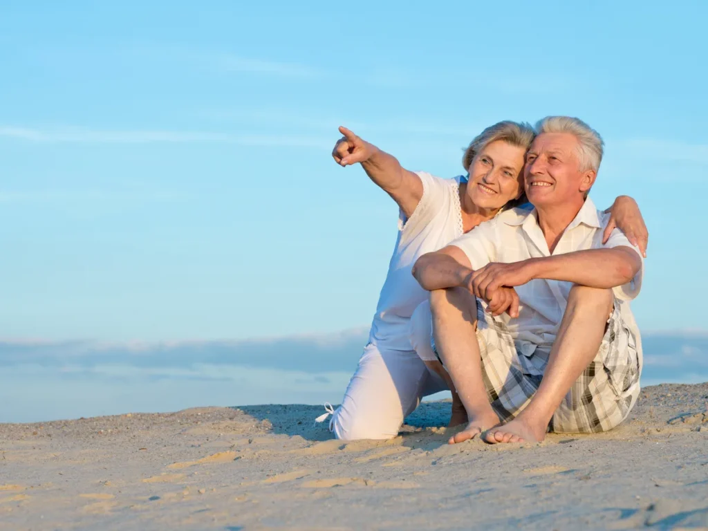 Older couple sitting on a sunny beach and pointing at the sea